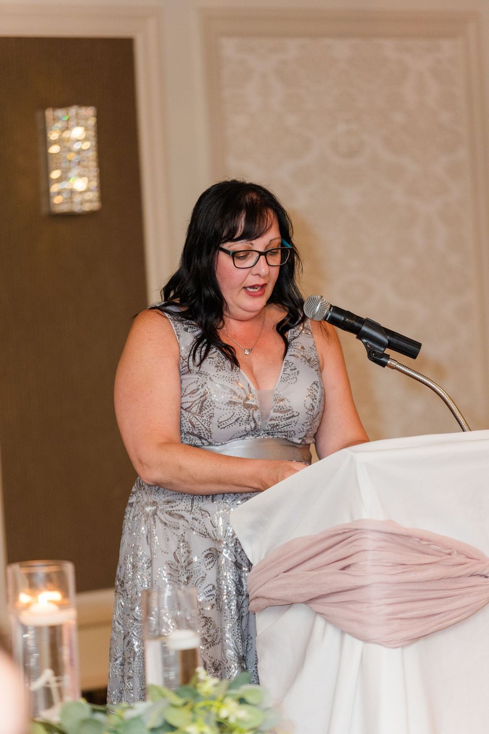 Bridesmaid in silver grey dress gives emotional speech at wedding reception podium with candles and decor visible.