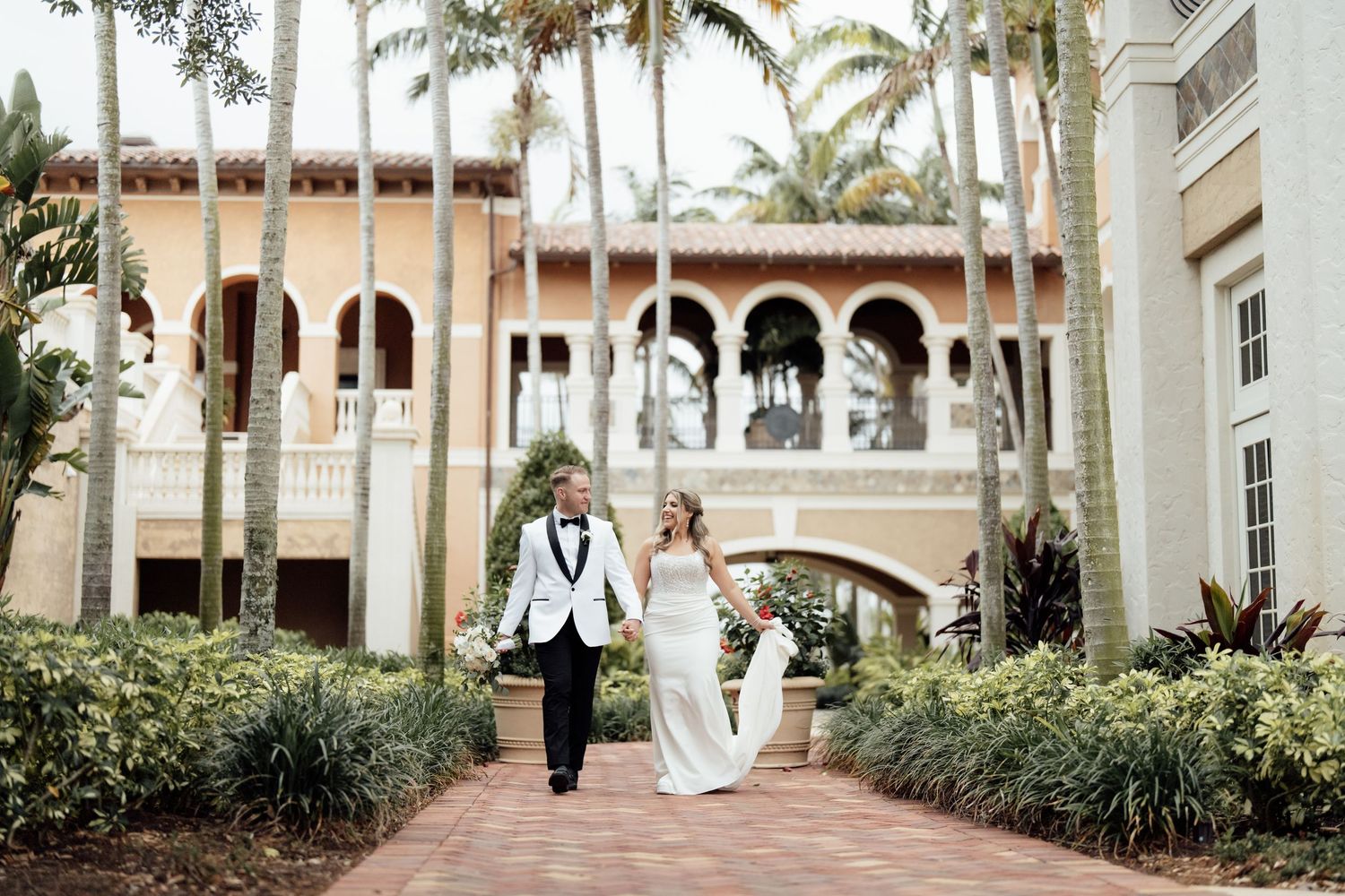 Newlyweds walk down a palm-lined path outside an elegant Mediterranean-style venue on their wedding day.