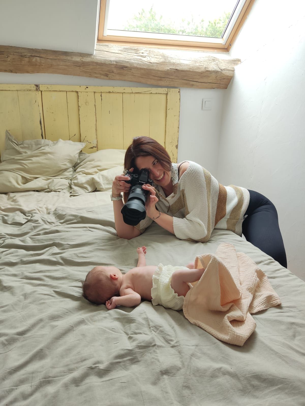 A photographer takes pictures of a baby lying on a bed under a skylight window in an attic bedroom.