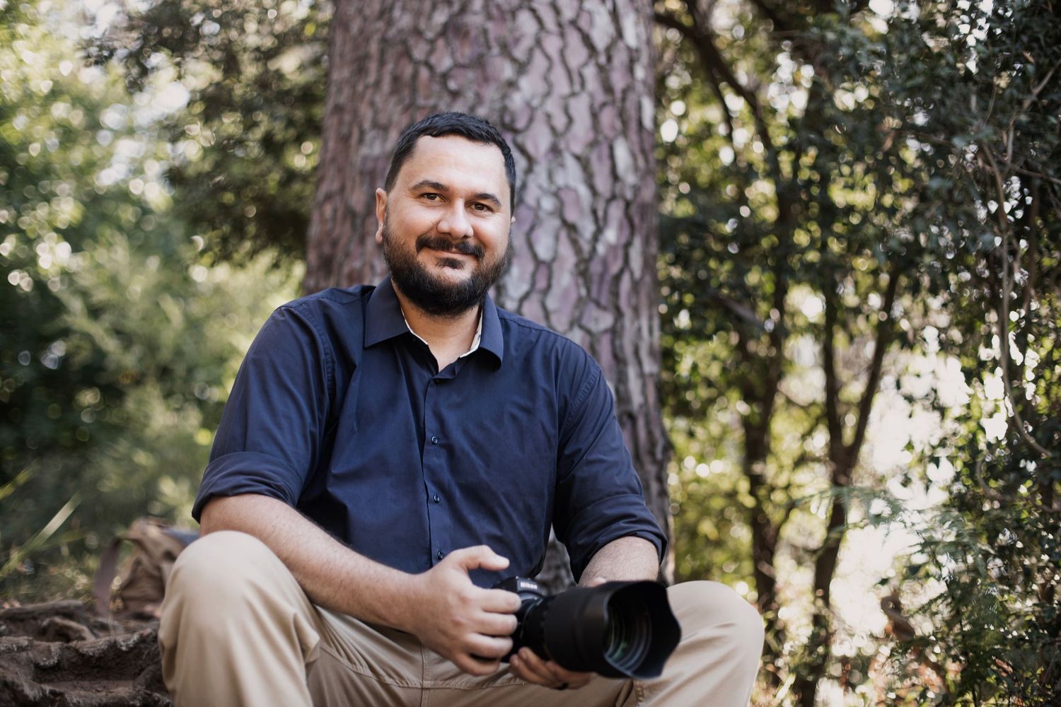 A smiling person in a navy blue shirt sits outdoors beside a pine tree in a natural setting.