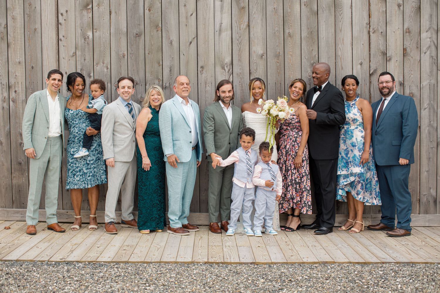 A group of wedding guests pose together in formal attire against a rustic wooden wall backdrop.
