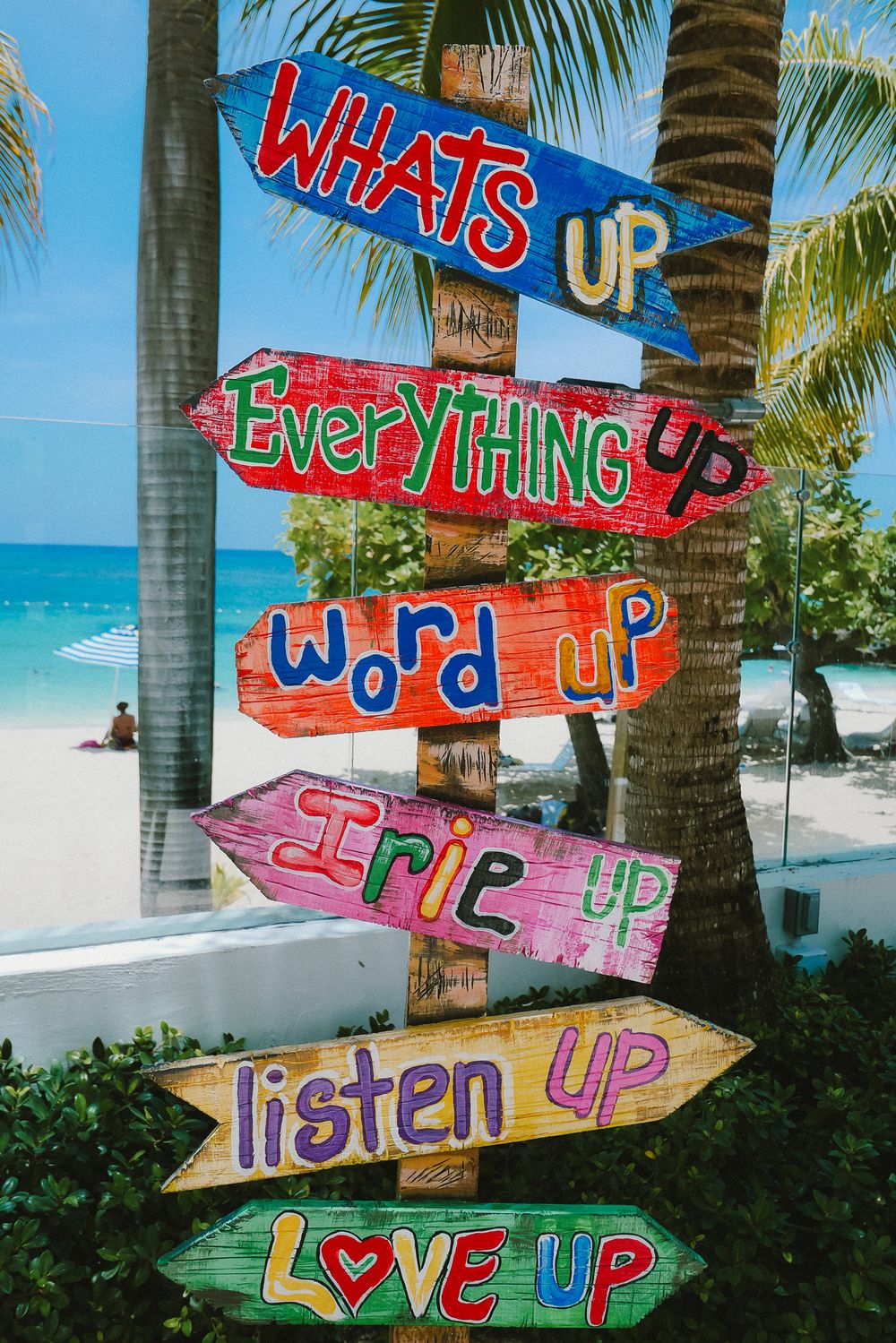 Colorful beach direction signs with inspirational words painted on driftwood against a tropical ocean and palm tree backdrop.