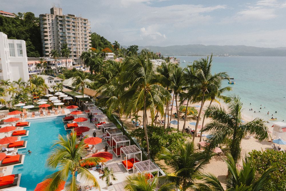 Aerial view of a luxury beachfront resort with palm trees, swimming pools, and red umbrellas along a turquoise coastline.