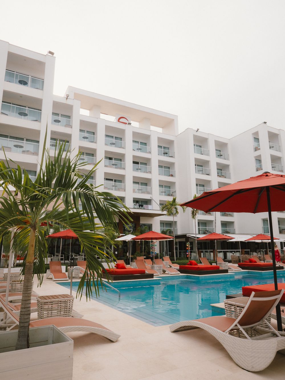 Luxury hotel pool area featuring white modern architecture, red umbrellas, and curved sun loungers surrounded by palm trees.