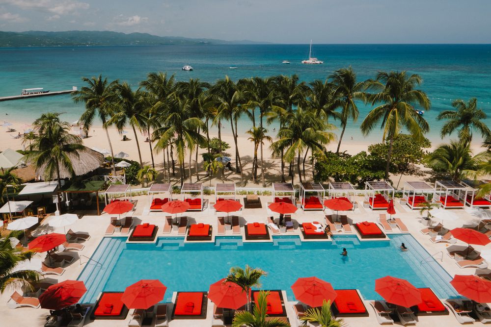 Stunning beachfront resort with vibrant red lounge chairs overlooking turquoise waters and swaying palm trees.