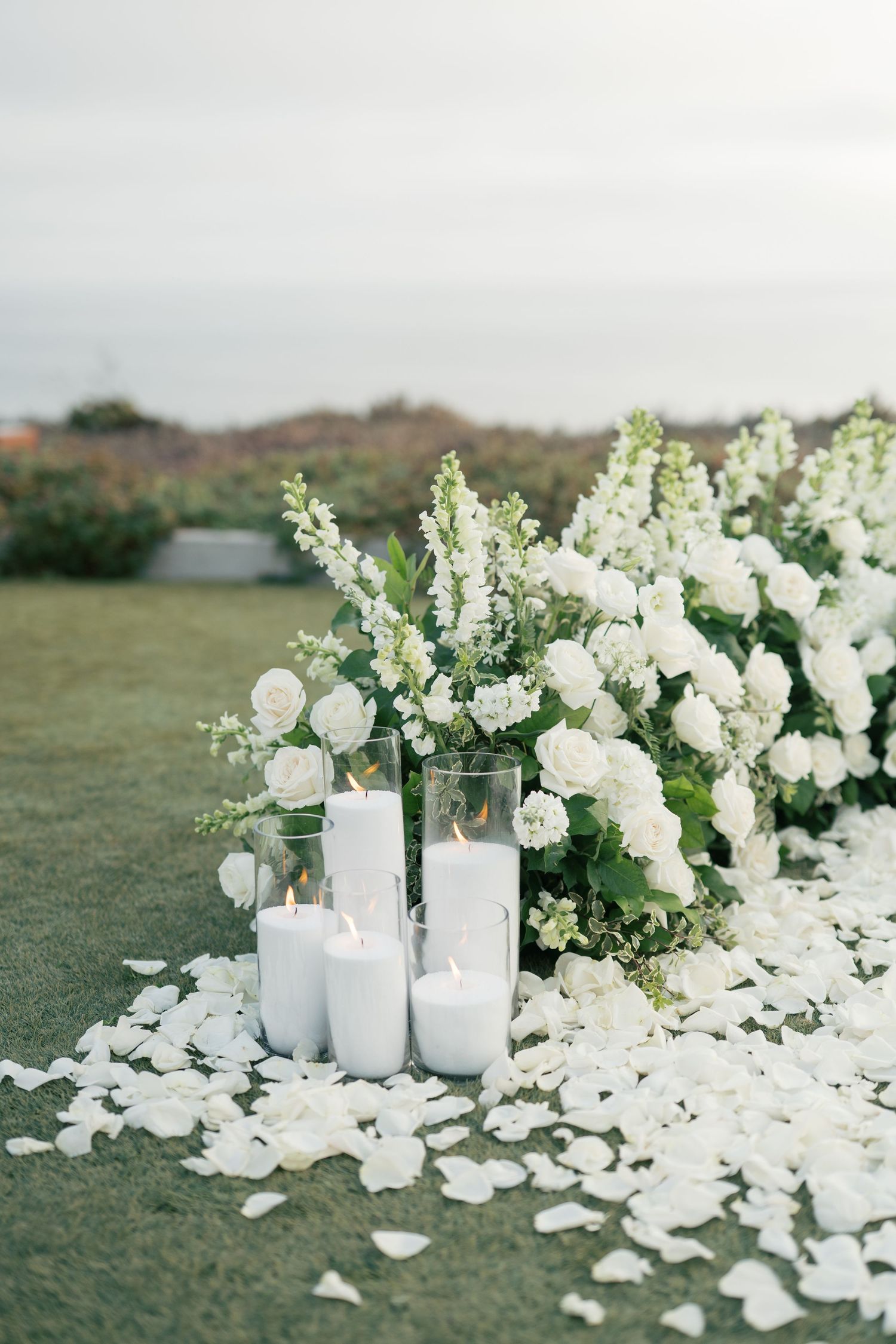 White floral arrangements with pillar candles and scattered petals create an elegant outdoor wedding display on grassy terrain.