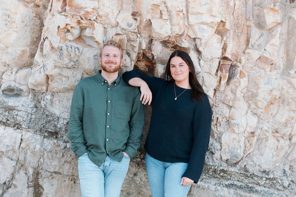A couple poses together against a rustic stone wall backdrop in coordinated green and black outfits.
