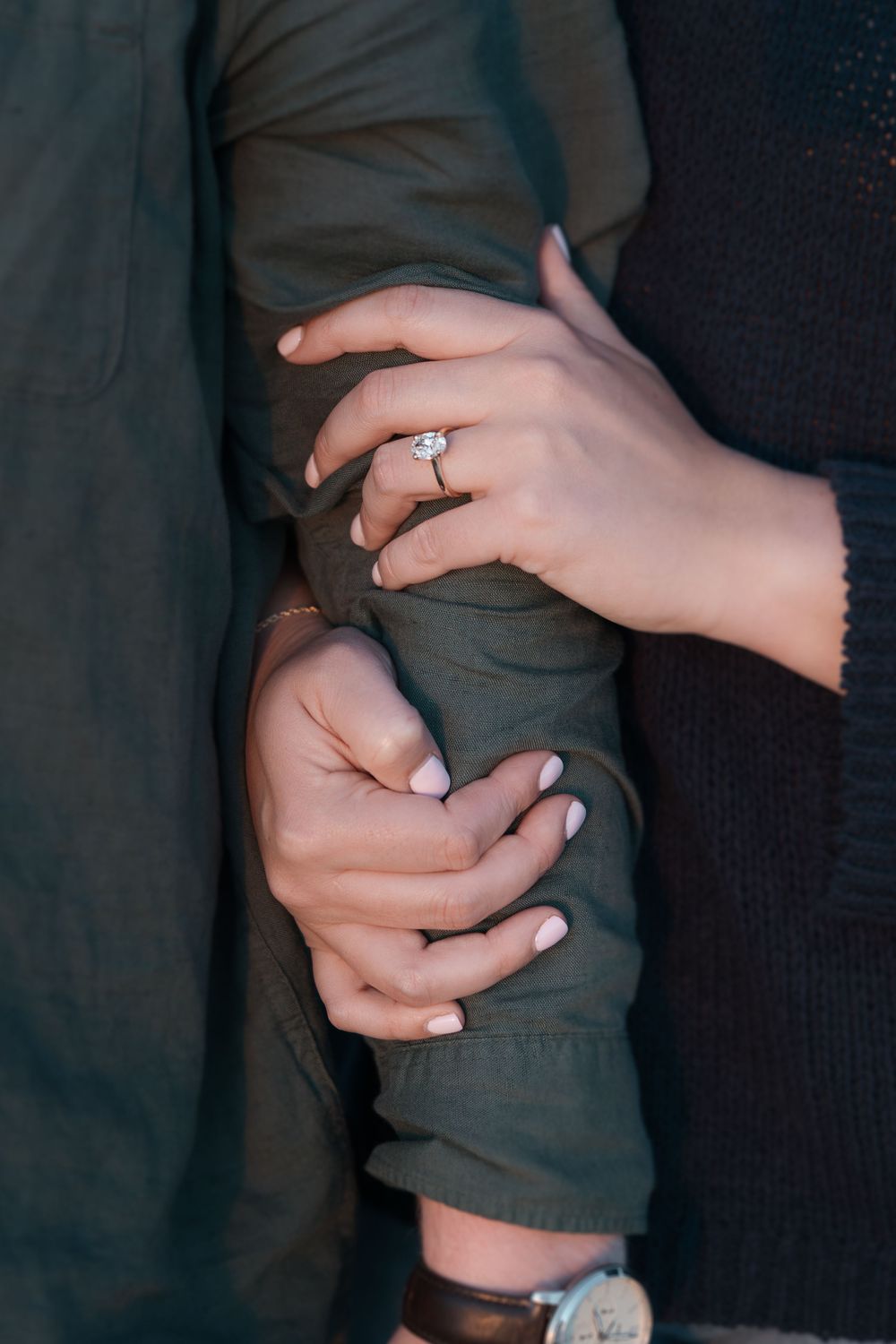Close-up of hands showing off an engagement ring against dark clothing.