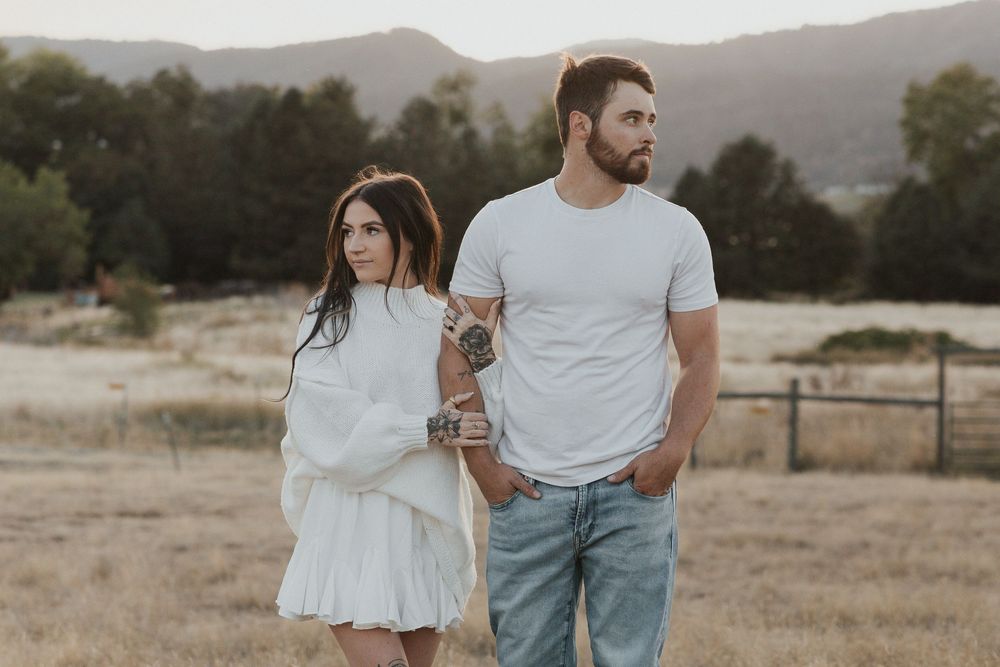A couple in casual attire walks together in a scenic field with mountains in the background.