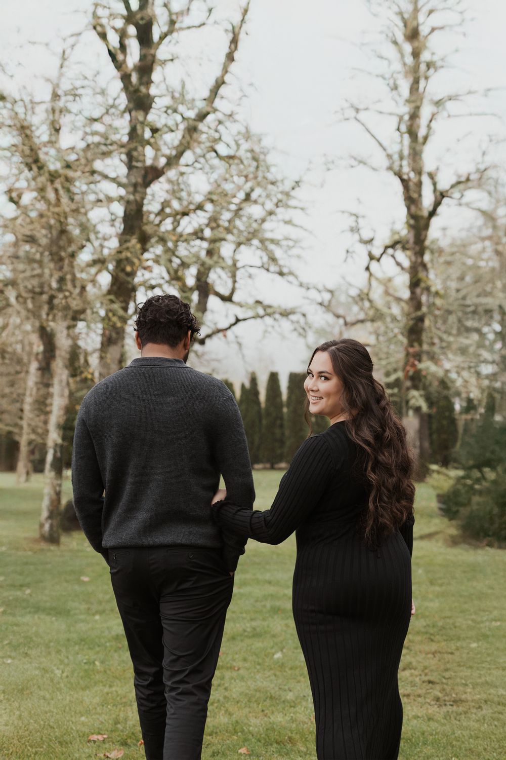 A couple in black attire walks together through a scenic park with bare trees in the background.
