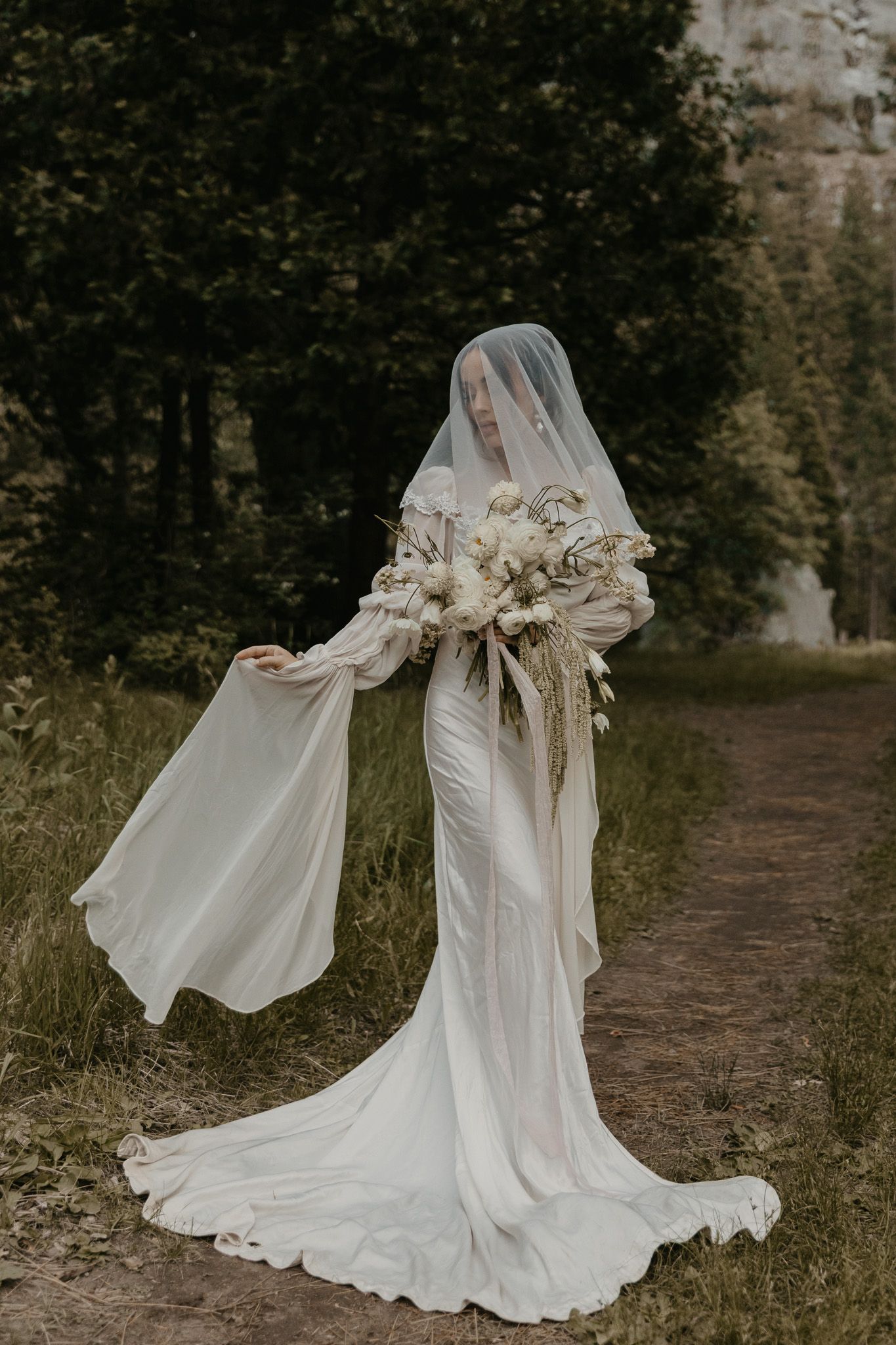 Flowing white wedding dress with translucent bell sleeves captured in motion against a natural outdoor backdrop.