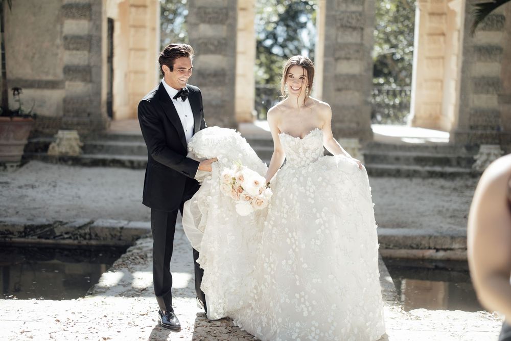 Wedding couple in elegant attire walking together through an ornate stone colonnade.