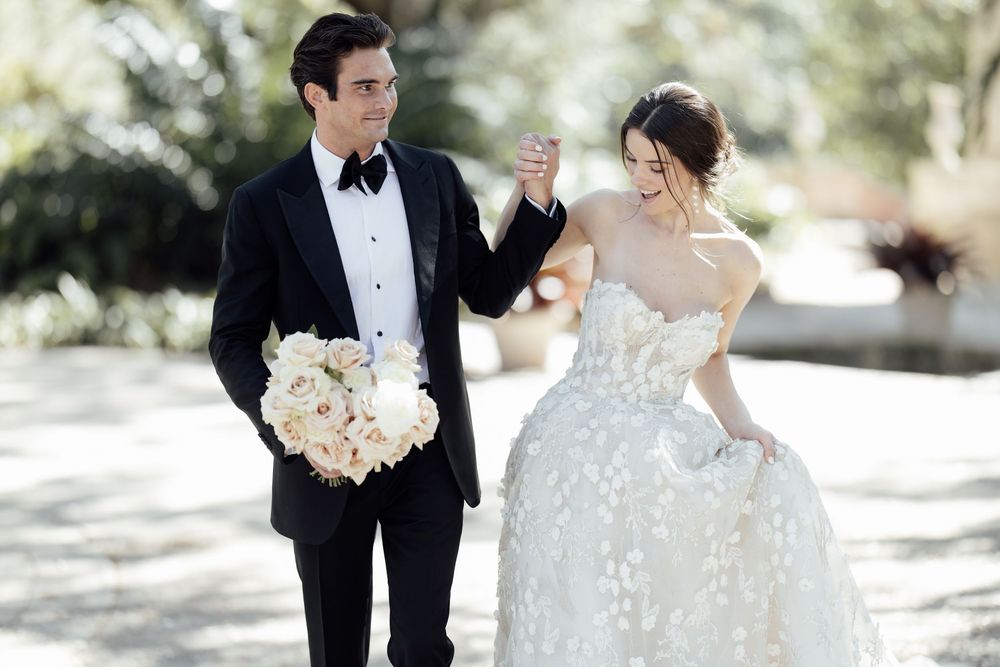 Newlyweds share a playful moment outdoors with white floral bouquet and elegant wedding attire.