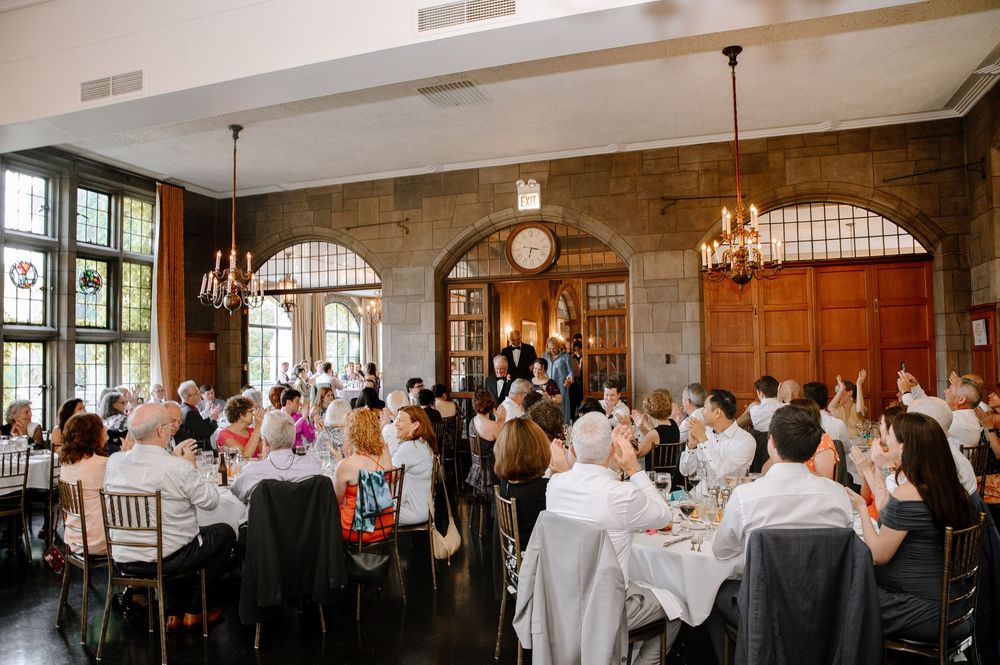 Elegant wedding reception in historic stone hall with arched windows and chandeliers as guests dine at round tables.