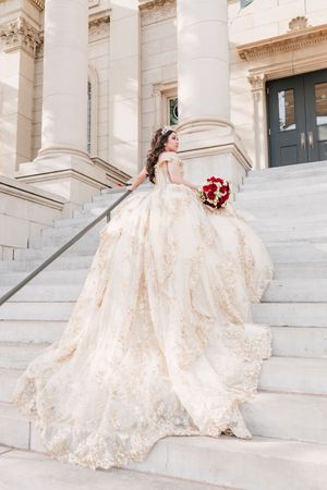 A bride in an ornate champagne lace gown sits gracefully on white marble steps of a grand classical building.