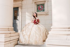 A bride in a champagne-colored ballgown poses elegantly on marble steps of a classic architecture building.