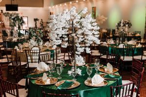 A wedding reception venue decorated with warm lighting, dark wooden chairs, and emerald green tablecloths.