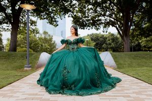 A person wearing a flowing emerald green quinceañera dress twirls on a stone pathway in an outdoor garden setting.
