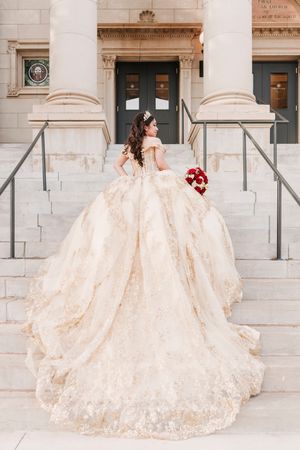 A bride in a champagne-colored ballgown with floral lace details poses against a white stone building.