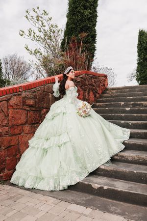 A person in a mint green ball gown poses elegantly on stone steps against a backdrop of brick walls and greenery.