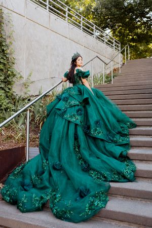 The flowing emerald green gown with delicate floral embellishments cascades dramatically down concrete steps outdoors.