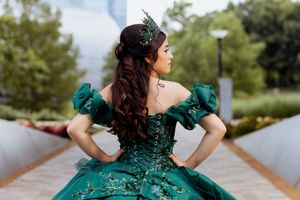Woman in emerald green formal dress poses elegantly outdoors near flowering purple bushes.