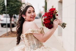 A bride in a champagne-colored gown holds a vibrant red rose bouquet while standing outdoors.