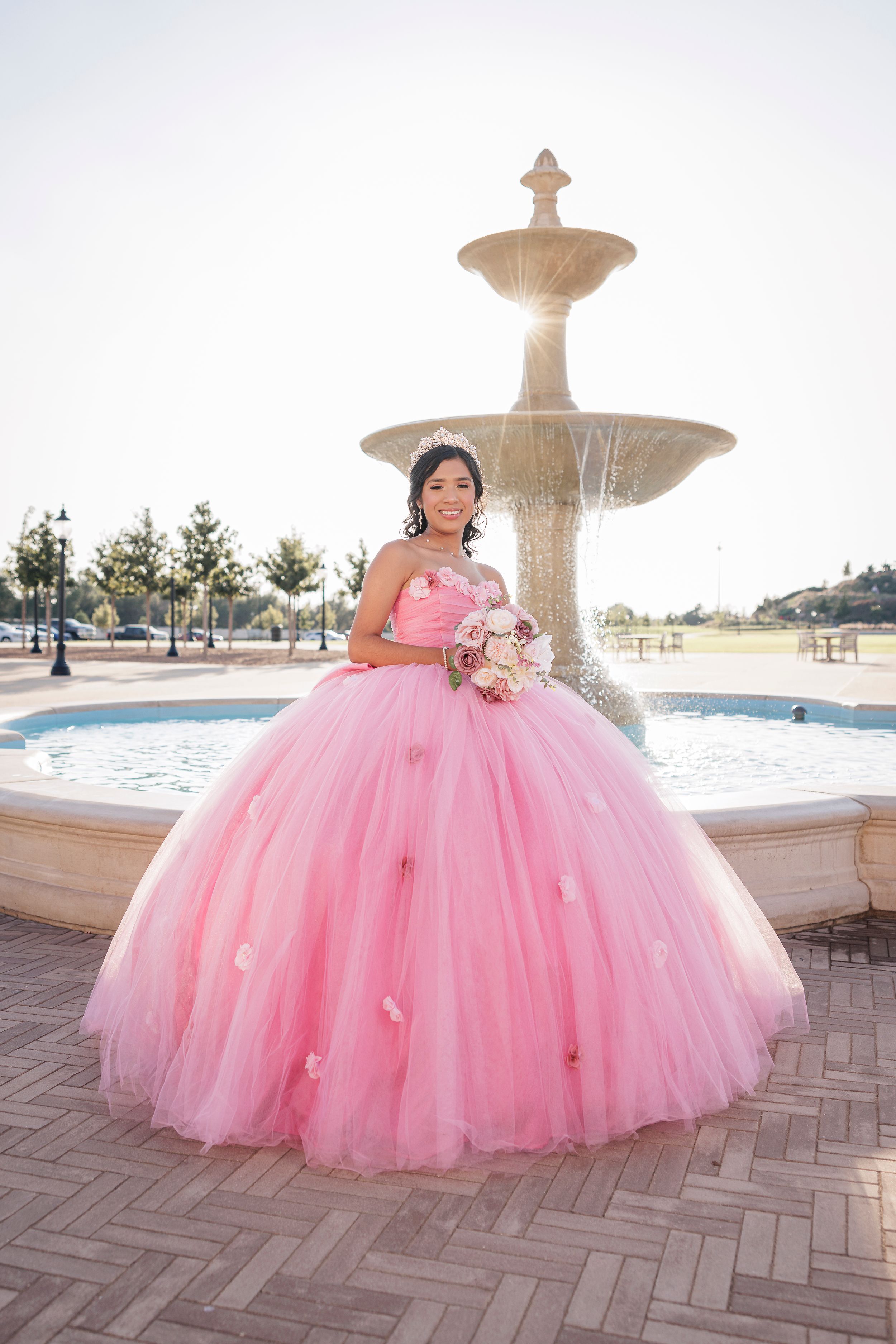 Stunning pink quinceañera ball gown with full tulle skirt photographed at sunset by a decorative fountain.