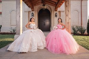 Two elegant ball gowns, one in white and one in pink, displayed against an ornate architectural backdrop.