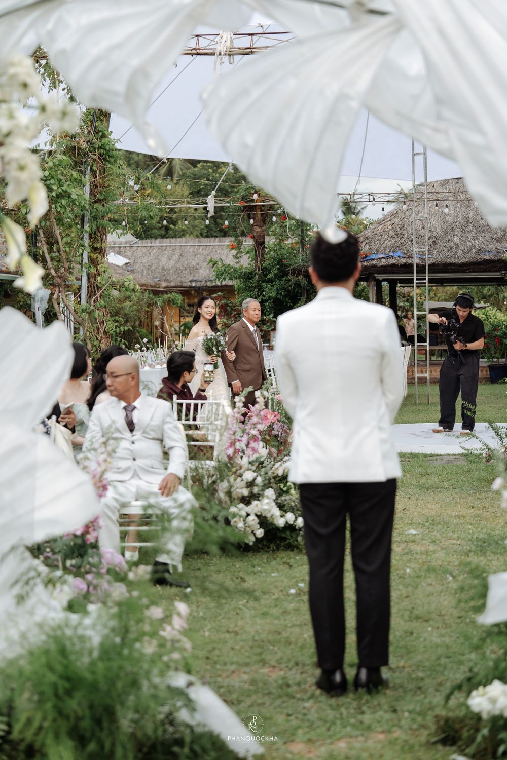 Groom in white suit awaits bride walking down the aisle at elegant outdoor garden wedding.