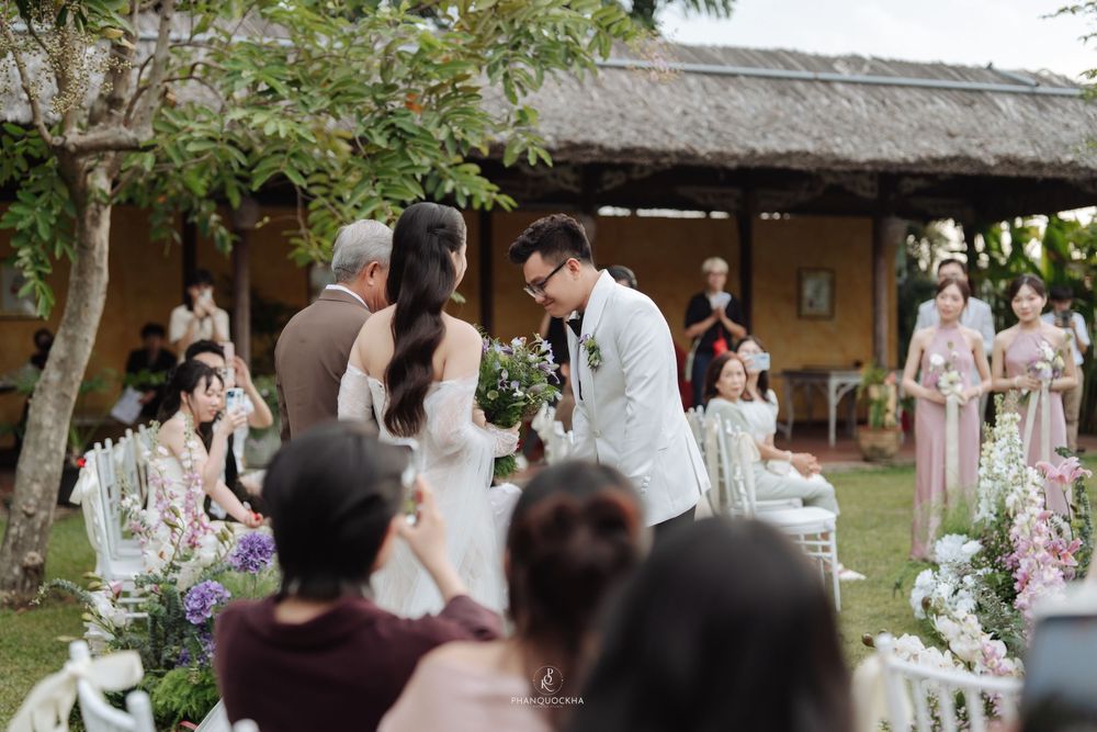 Groom in white suit greets bride during outdoor garden wedding ceremony with floral decorations.