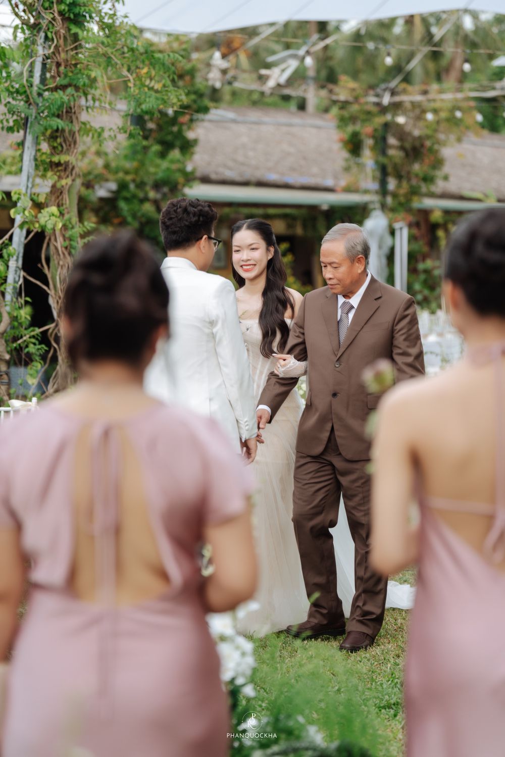 Bride in white gown smiles as father escorts her to groom at outdoor wedding ceremony.