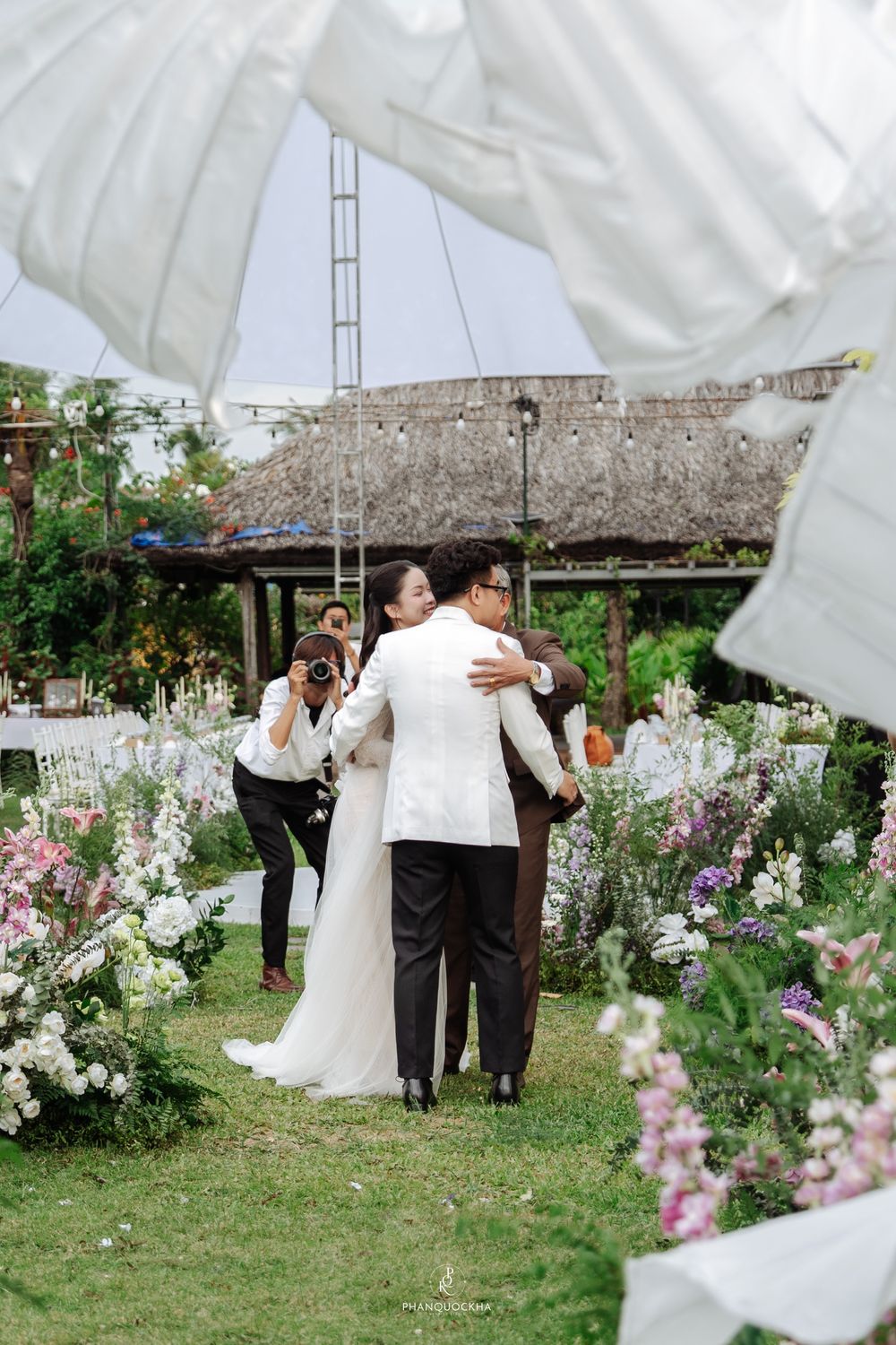 Bride and groom share a warm embrace on the floral-lined aisle of an elegant outdoor garden wedding ceremony.