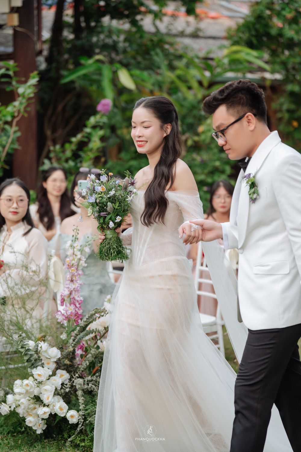 Smiling bride in off-shoulder white gown holds wildflower bouquet while groom in white tuxedo escorts her down aisle.