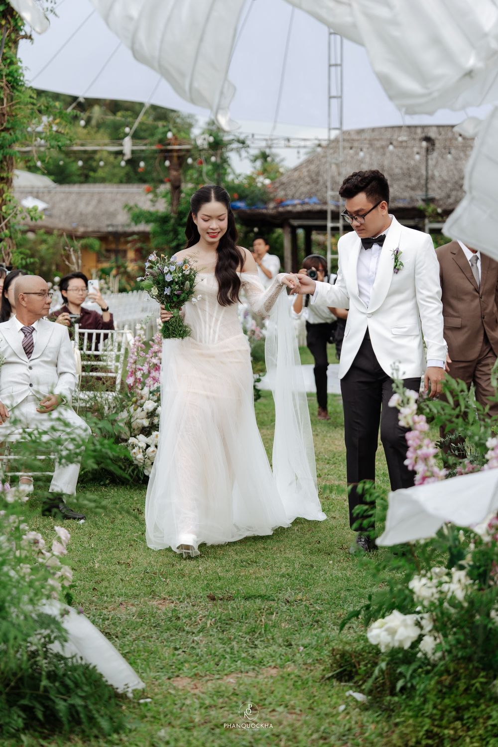 Bride and groom walk down garden aisle lined with white chairs and floral arrangements under billowing white sculptures.