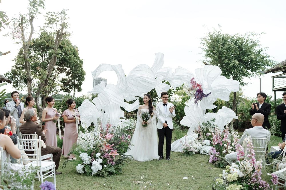 Wedding couple celebrates at outdoor ceremony beneath large white fabric sculptures with guests seated on both sides.