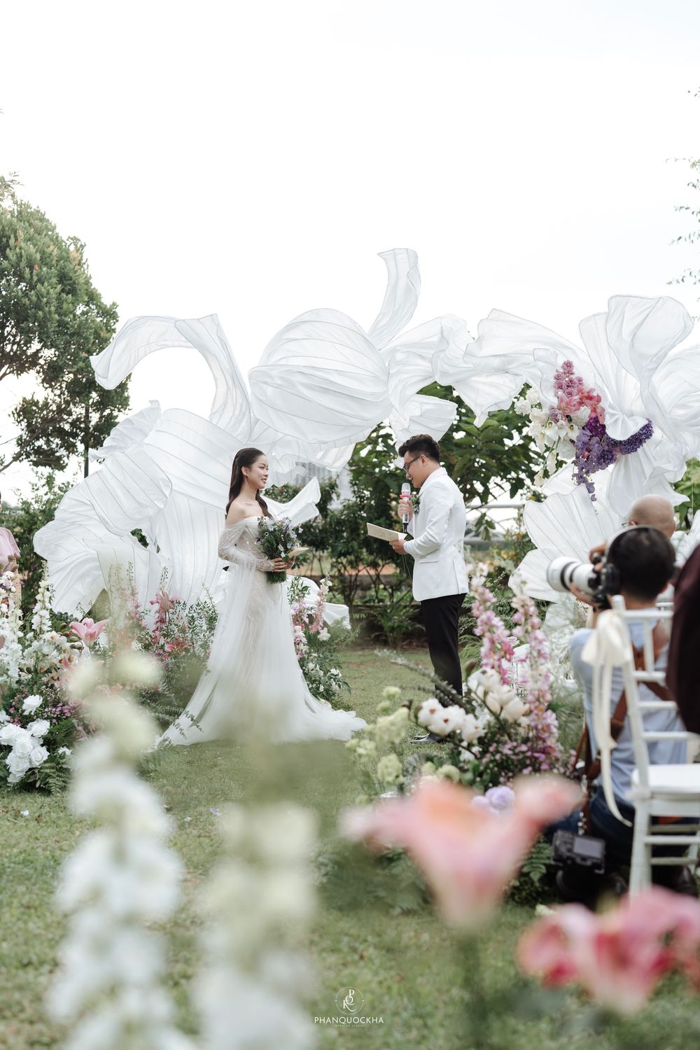 Bride and groom exchange vows beneath dramatic white fabric arch with colorful floral arrangements at outdoor ceremony.