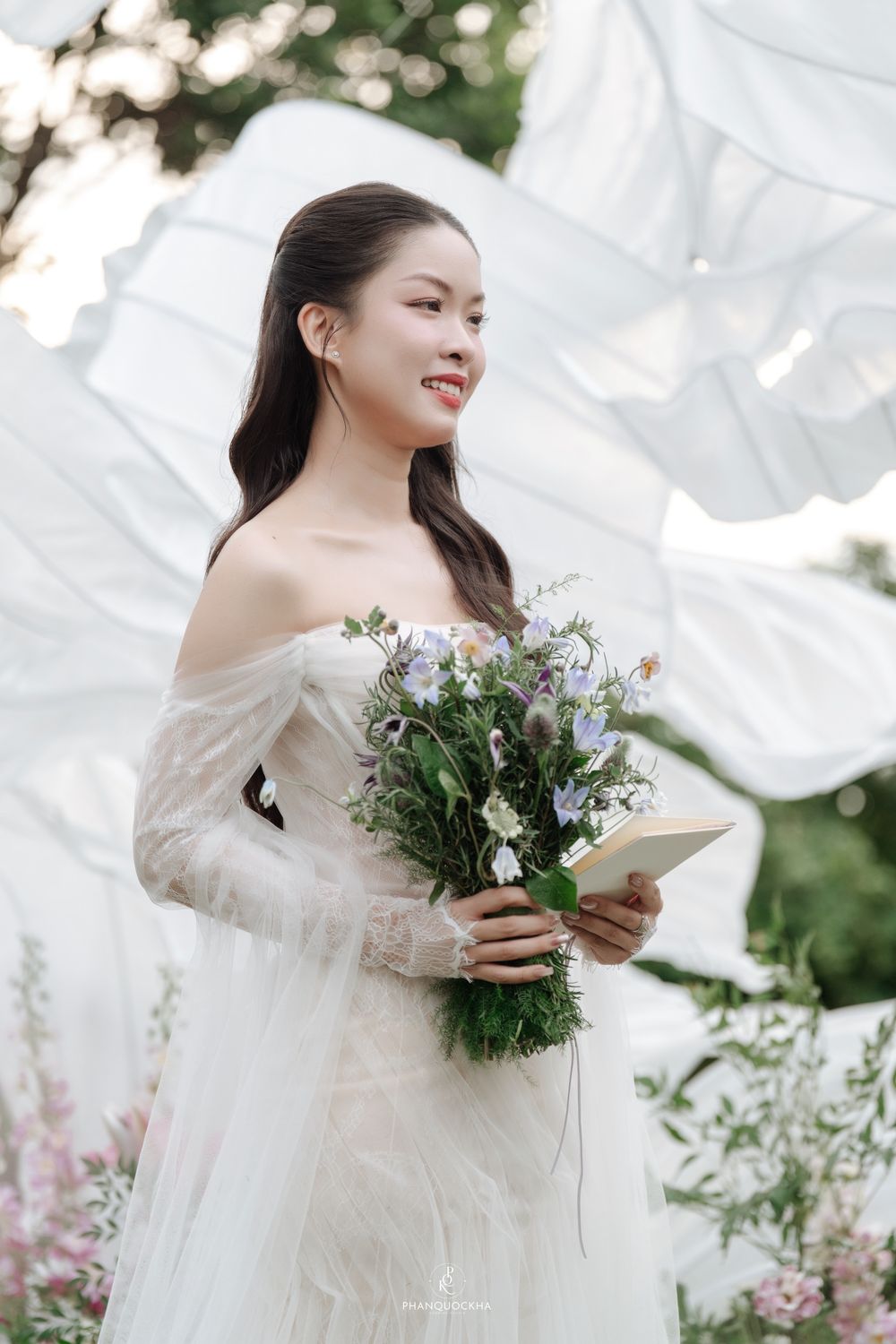 Radiant bride in off-shoulder white lace gown holds wildflower bouquet and card near billowing white fabric backdrop.