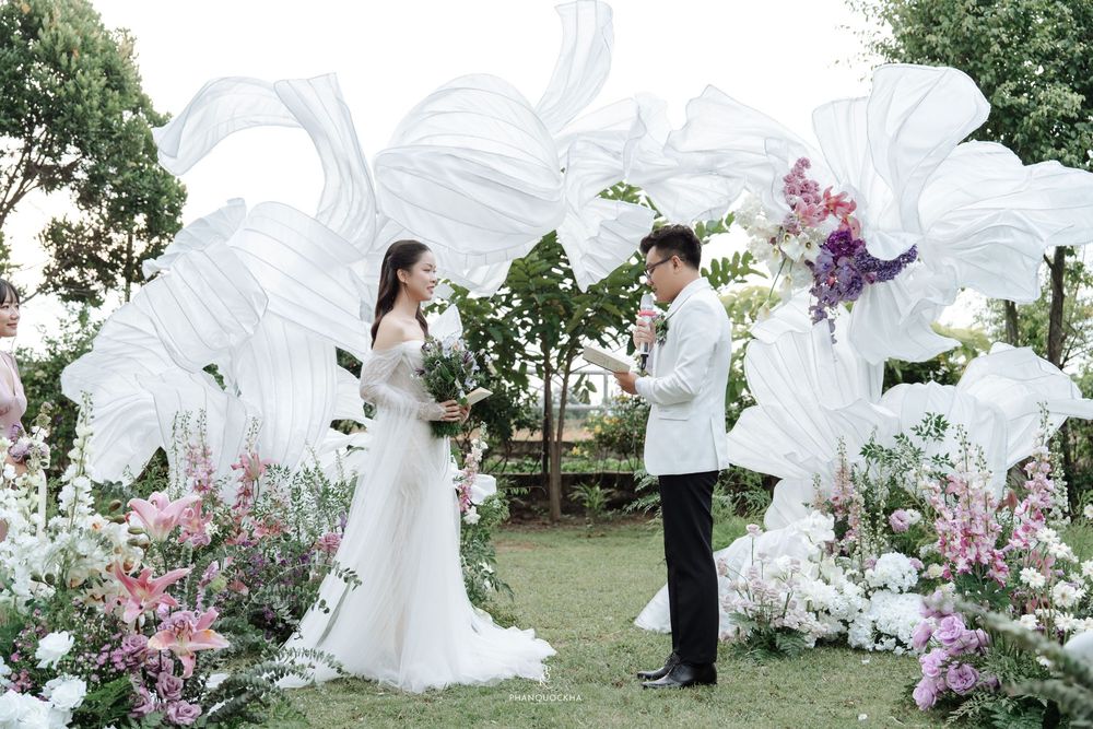 Bride and groom face each other exchanging vows beneath white fabric arch adorned with purple and white flowers.