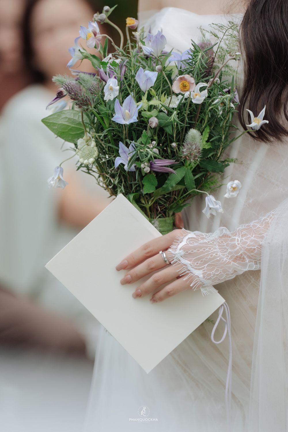Close-up of bride holding wildflower bouquet with blue blooms and cream envelope with lace wedding dress sleeve detail.