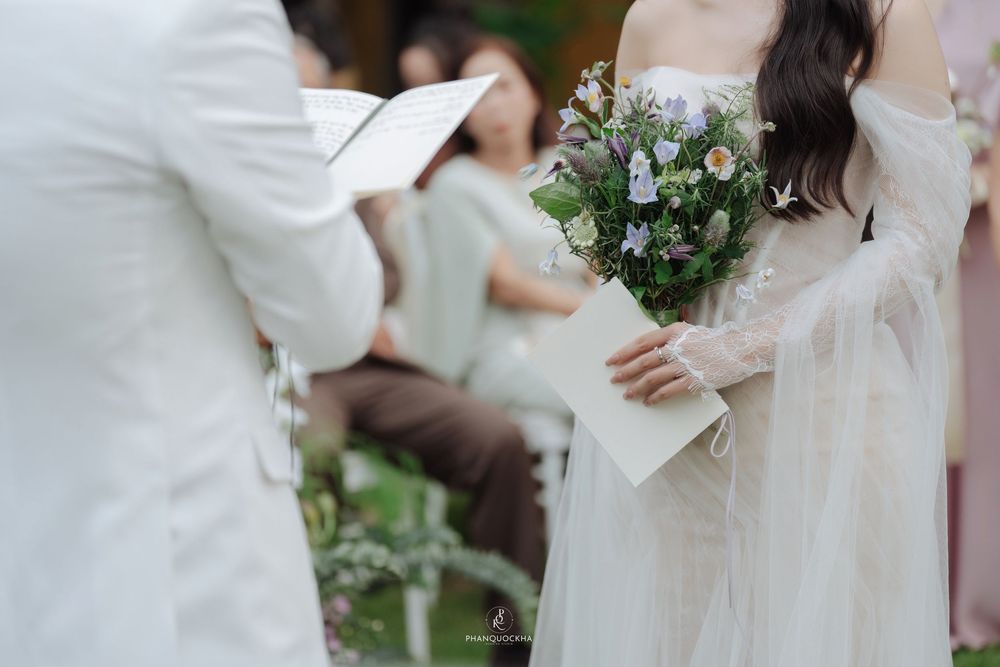 Bride in white lace gown holding wildflower bouquet during outdoor wedding ceremony.