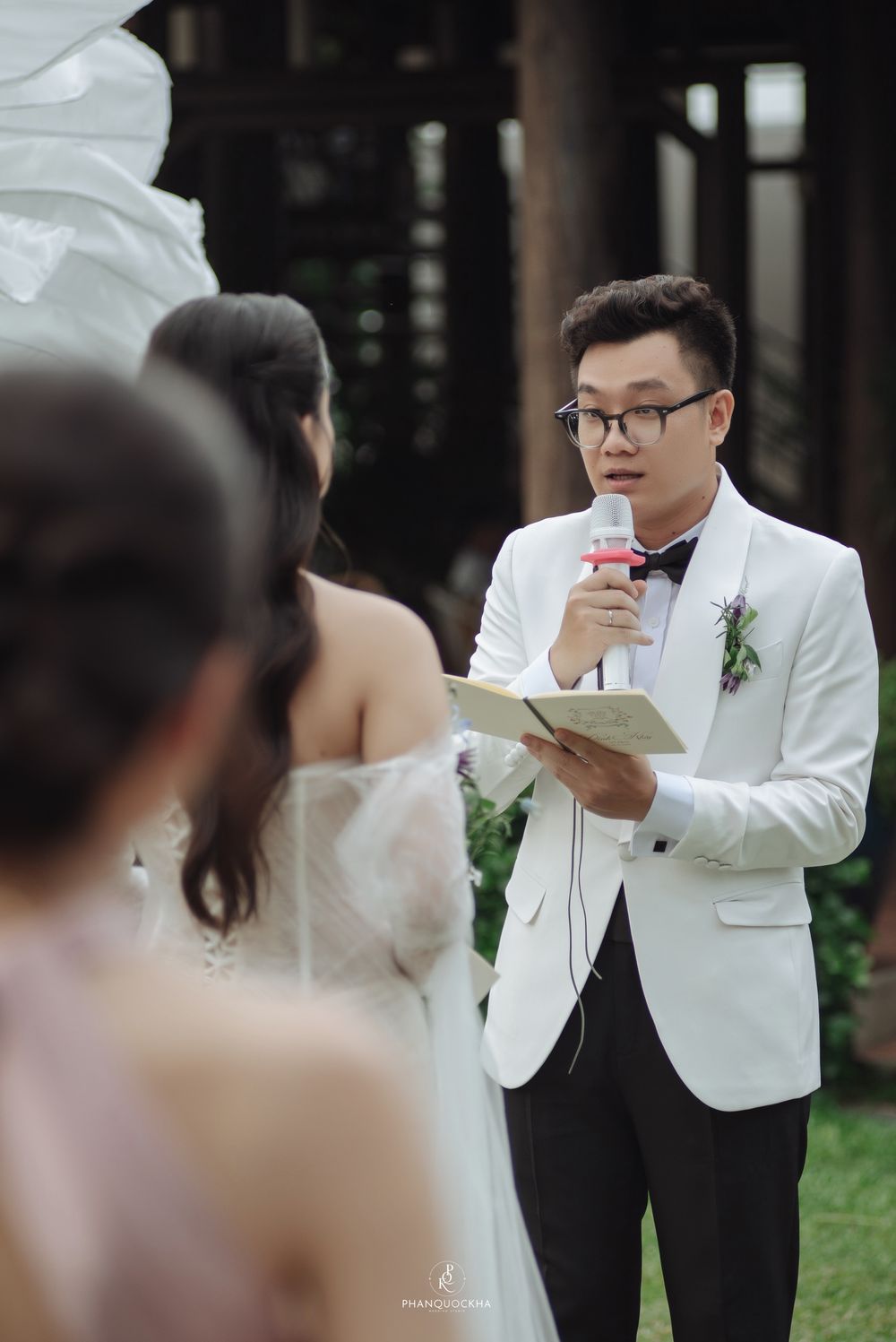 Groom in white tuxedo reading vows into microphone at outdoor wedding ceremony.