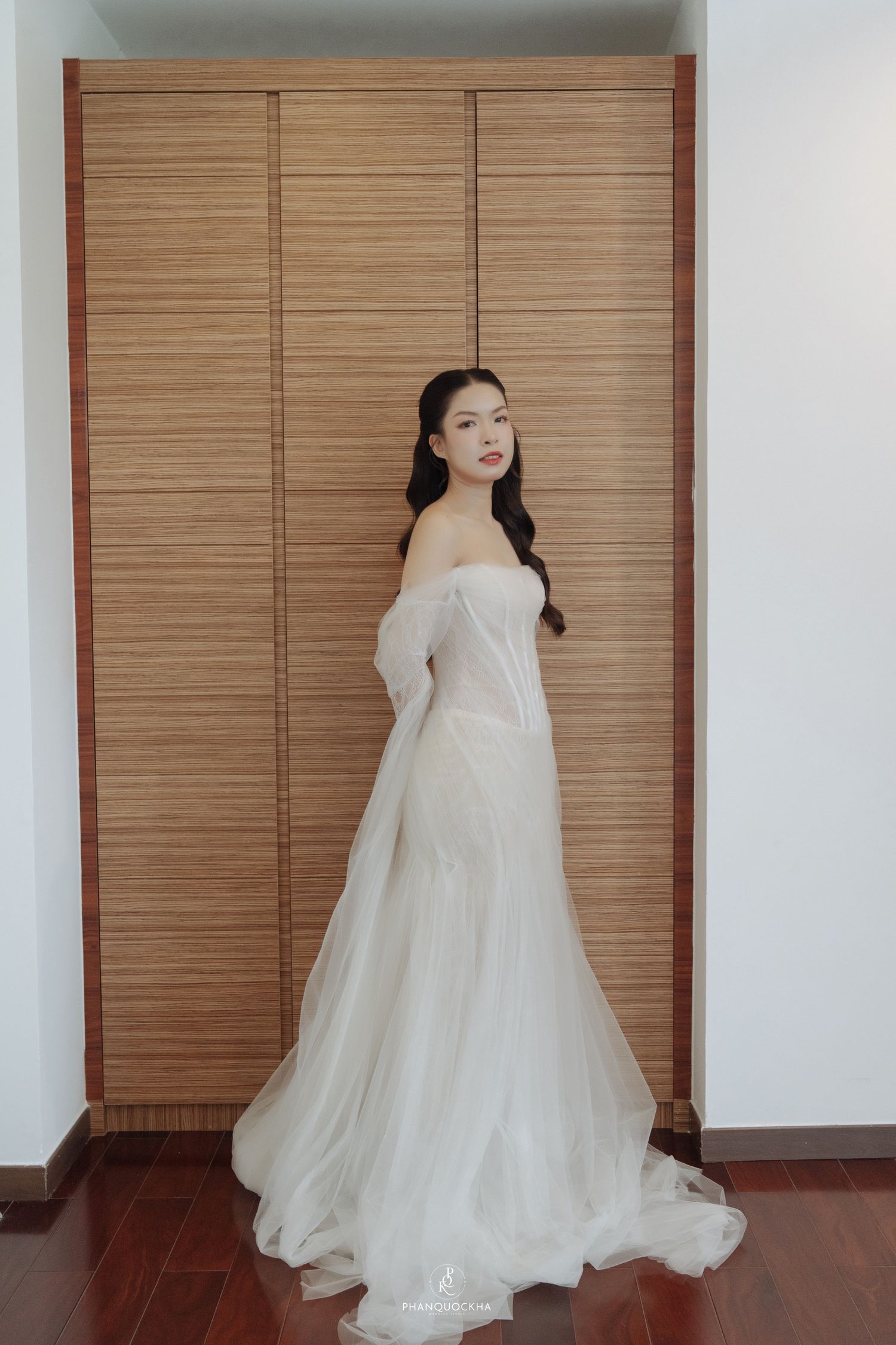 Beautiful Asian bride in off-shoulder white mermaid wedding gown posing by wooden wardrobe.