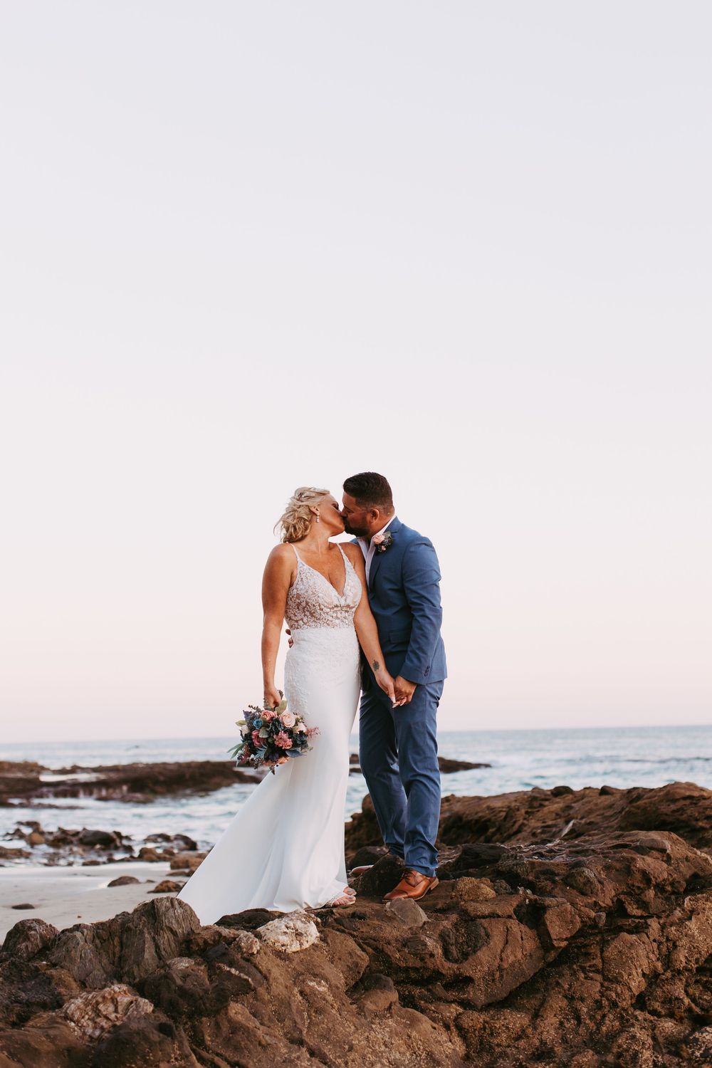 Newlywed couple shares romantic moment on rocky coastal beach at sunset with ocean waves in Laguna Beach, California.