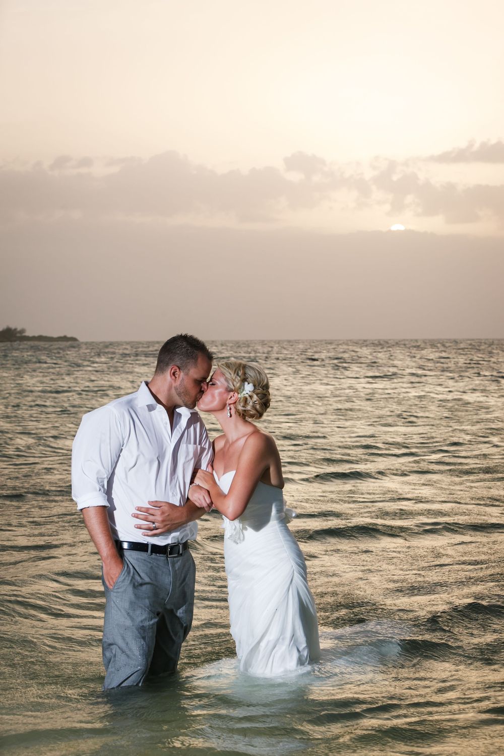 A romantic couple shares a kiss while standing in ocean waters at sunset, creating a dreamy beach scene.