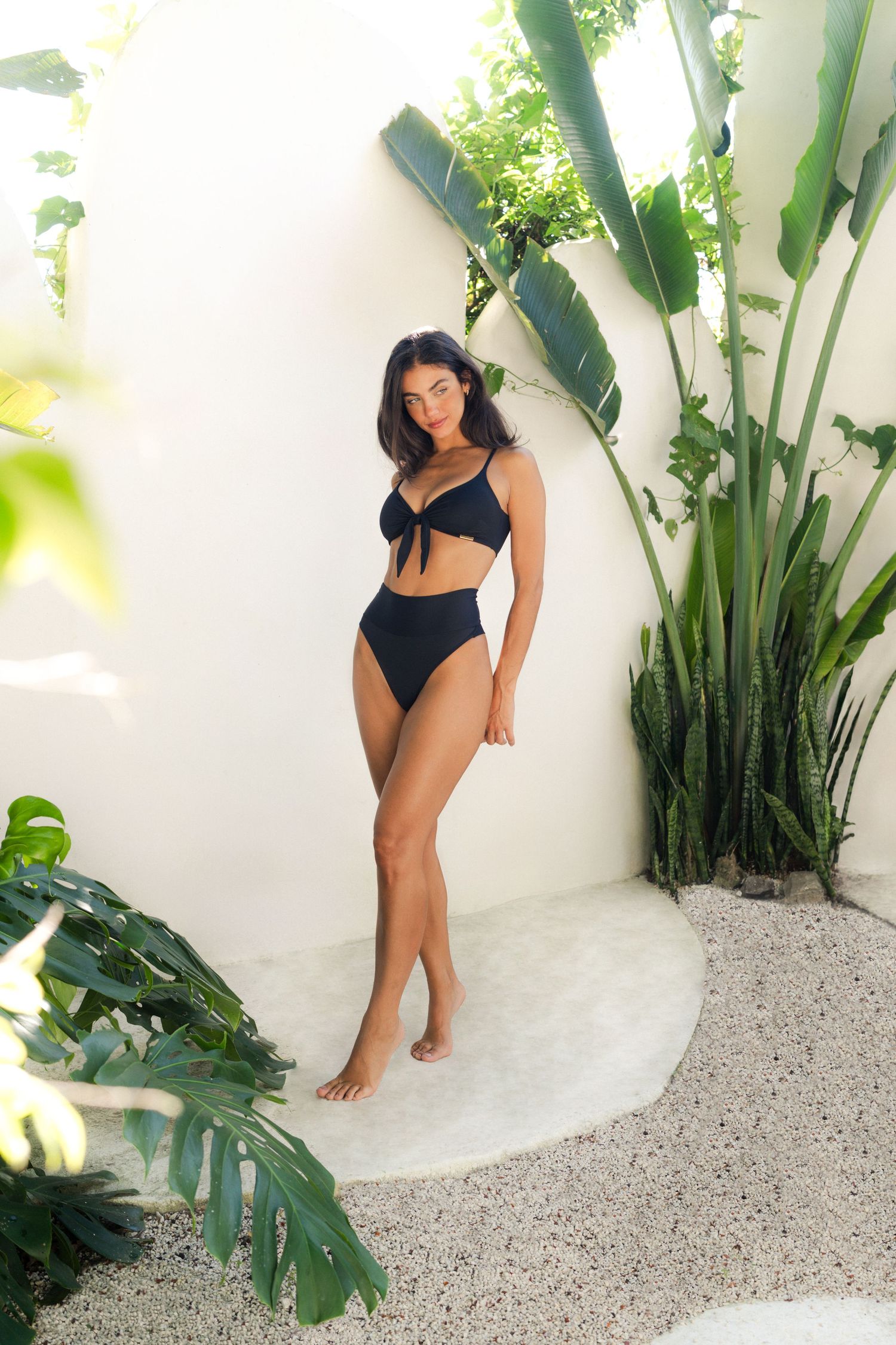Figure in a black swimsuit poses elegantly against a tropical backdrop with large palm leaves and white walls.