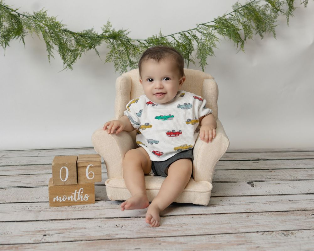 A small child sits in a white chair beneath evergreen garland with a wooden milestone block marking 6 months.