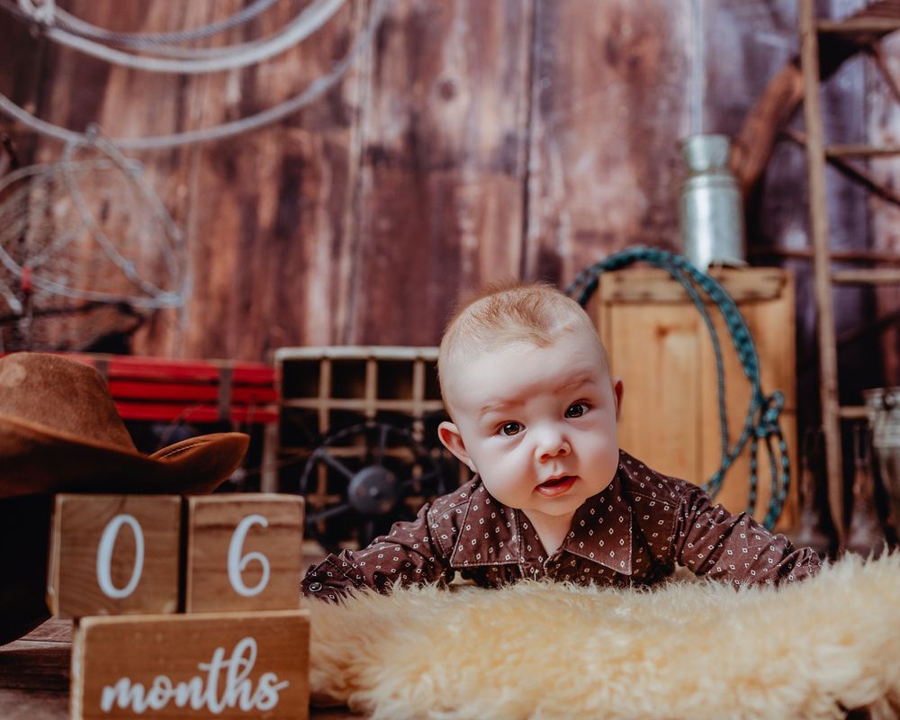 Baby wearing flowered outfit lies on fuzzy rug next to wooden 6-month milestone marker in rustic setting.