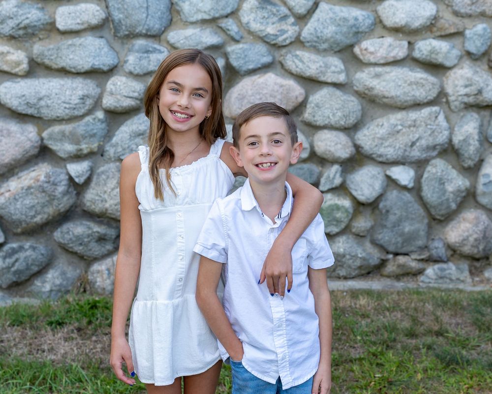 Siblings in white outfits pose together against a rustic stone wall background during a fall family photo session in Sudbury, MA.