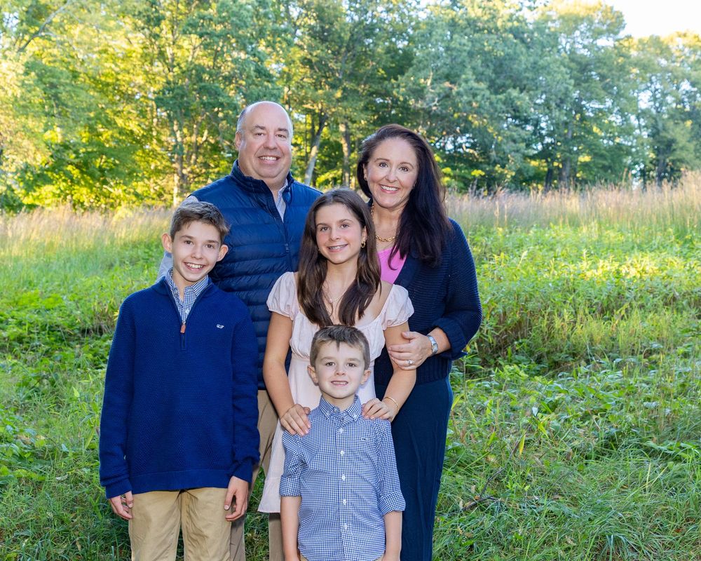 A family poses together outdoors in a sunlit field with green grass and trees in the background during a family photography session Concord.